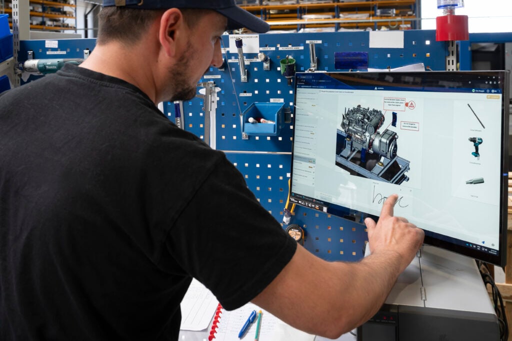 Un operario de e-power usa una pantalla táctil para seguir instrucciones de trabajo digitales destinadas al montaje de una unidad de potencia. La pantalla muestra una imagen 3D detallada de un motor con anotaciones y referencias a herramientas, impulsada por la plataforma Azumuta. Al fondo, las herramientas están cuidadosamente organizadas en un panel perforado azul y sobre la estación de trabajo se ven un cuaderno y bolígrafos.
